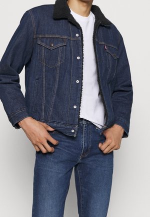 Mand iført mørkeblå denimjakke med sort fleecekrave, hvid t-shirt og blå jeans, poserer med hænderne nær lommerne.