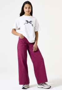 Camiseta blanca de algodón con un gráfico de lazo gris, combinada con pantalones anchos color burdeos y zapatillas deportivas plateadas. Diseño simple, ajuste casual.