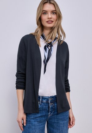 Donna che indossa un cardigan nero aperto, una maglietta bianca, jeans blu e una sciarpa a righe intorno al collo, in piedi davanti a uno sfondo chiaro e uniforme.