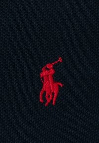 Námořnická pletenina obsahuje červeně vyšitý logo koně a jezdce, který drží trumpetu, umístěné uprostřed.