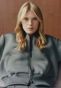 Felpa grigia con zip, polsini e orlo a coste, dotata di due tasche laterali e una texture liscia, indossata da una persona con capelli lunghi.