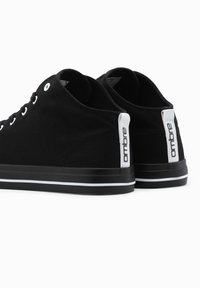 Zapatillas de lona negras con cordones blancos, punta redondeada, rayas blancas contrastantes en la suela y una marca minimalista en el talón.