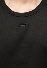 Κοντινό πλάνο μαύρης μπλούζας με λαιμόκοψη, με ένα μικρό κεντημένο διπλό κύμα λογότυπο στο κέντρο κάτω από τη λαιμόκοψη.