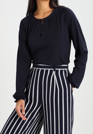 Frau trägt ein marineblaues Langarmshirt mit Knöpfen und hoch taillierte marineblaue Hose mit vertikalen weißen Streifen, Hand in der Tasche.
