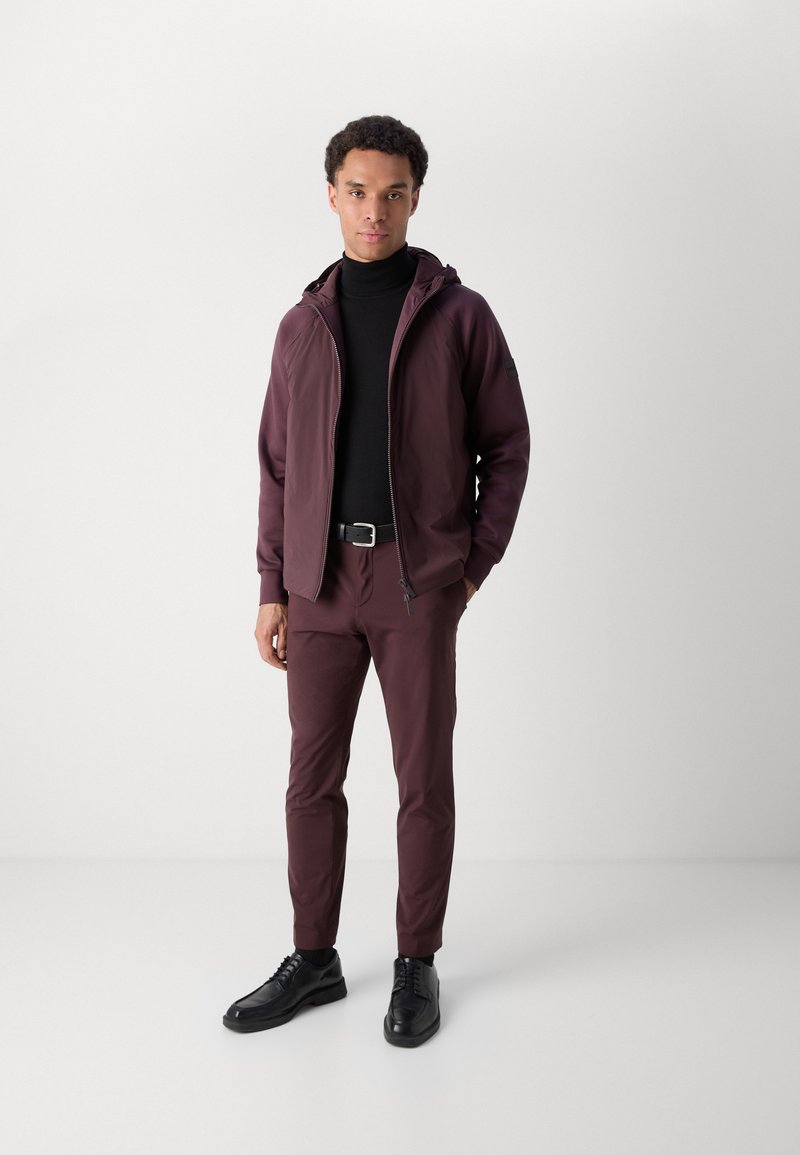 Bordeauxfarbene Zip-Up-Jacke mit Kapuze, kombiniert mit einem schwarzen Rollkragenpullover und passenden bordeauxfarbenen Hosen. Schwarze Schnürschuhe runden das Outfit ab.