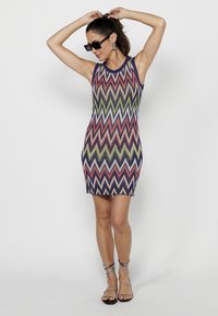 Rochie bodycon tricotată cu model zigzag multicolor; fără mâneci, gât rotund și tiv contrasting. Asortată cu sandale negre cu curea.