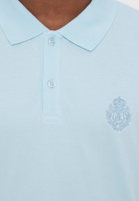 Svetlo modra polo majica s klasičnim ovratnikom, tremi gumbi in teksturirano tkanino. Na levi prsni strani ima vezen grb.