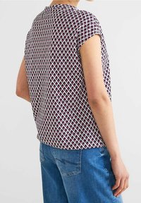 Top a maniche corte, stampato in bianco con forme a diamante in viola e rosa. Tessuto morbido con una vestibilità rilassata, abbinato a jeans blu.