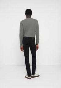 Maglia grigia a maniche lunghe, texture a coste, abbinata a jeans neri aderenti e scarpe bianche con suola marrone. Vista posteriore su sfondo bianco.