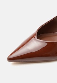 Καφέ γυναικείο παπούτσι με μυτερή μύτη, γυαλιστερό και λείο φινίρισμα, με χαμηλή κοπή στο άνοιγμα, σε λευκό φόντο.