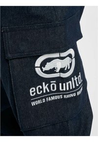 Dark denim cargo pants featuring a prominent embroidered logo of a rhino in a circular design, with "WORLD FAMOUS RHINO BRAND" text below.