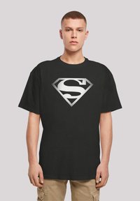 Must värviline T-särk mustast puuvillast, millel on suur valge Superman'i logo punktmustriga, ümmargune kaelus ja lõdvestatud istuvus, mudel seismas lihtsa tausta ees.