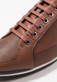 Baskets en cuir marron avec accents perforés, lacets plats, œillets métalliques et semelle en caoutchouc blanche. Le cuir texturé apporte des détails.