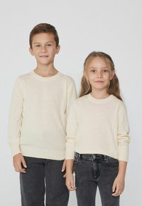 Cremefarbene gestrickte Pullover mit langen Ärmeln und gerippten Manschetten, kombiniert mit dunkelgrauen Jeans, getragen von zwei Kindern.