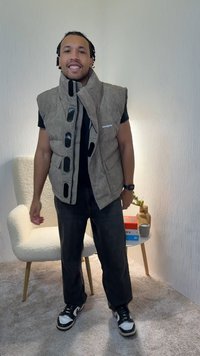 Καφέ φουσκωτό γιλέκο με ψηλό γιακά και μαύρα κουμπιά τύπου σκρατς. Φορεμένο πάνω από μαύρο πουκάμισο· συνδυασμένο με σκούρο παντελόνι και μαύρα-άσπρα αθλητικά παπούτσια.