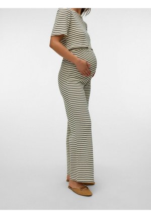 Gravid kvinde i beige og sortstribet matchende top og bukser, iført brune flade slip-on sko, hånd hvilende på maven.