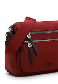 Červená nylonová crossbody taška s dvěma zipovými oddíly, hnědými koženými akcenty a nastavitelným textilním popruhem. Logo je umístěno na přední straně.