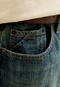 Τσέπη από τζιν με πορτοκαλί ραφές και μια μικρή μεταλλική ετικέτα που γράφει "EST.1990", φορεμένη με λευκό πουκάμισο και καφέ δερμάτινη ζώνη.