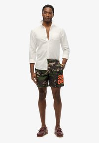 Chemise blanche à manches longues, short de bain camouflé vert et marron avec "SUPERDRY" en orange, chaussures marron, debout sur un fond neutre.