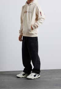 Felpa con cappuccio beige con tasca frontale e logo, abbinata a jeans neri larghi e sneakers bianche con dettagli blu navy.