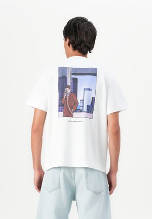 Man wearing white t-shirt with graphic of a suited man talking on a corded phone against a city skyline on the back, paired with light jeans.
