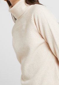 Kobieta ubrana w kremowy, prążkowany golf, pokazująca górną część torsu i detale dekoltu.