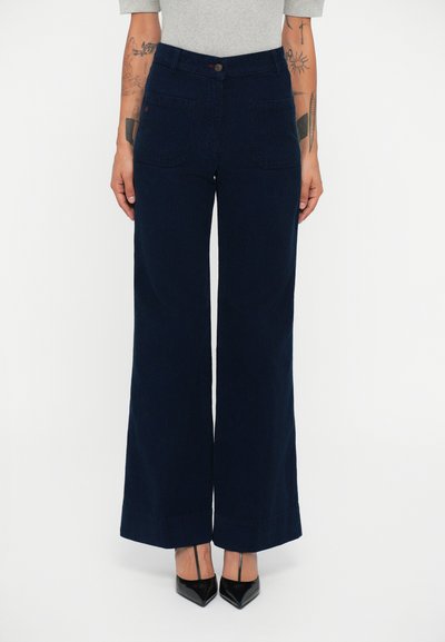 Jeans de pernera ancha, de color azul marino, hechos de tela tejida. Presentan una cintura alta, bolsillos frontales y cierre de botón. Se combinan con tacones puntiagudos negros.