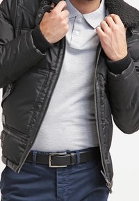 Doudoune en nylon noir avec une fermeture éclair avant, un col en peau lainée et des poignets en tricot ; portée sur un polo gris et un pantalon bleu, avec une ceinture noire.