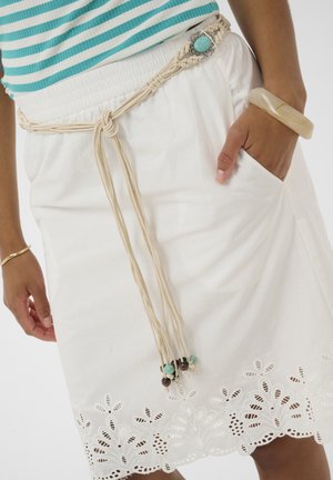 Kvinde iført hvid nederdel med blomsterbroderet kant, makramébælte med turkise sten, turkisstribet top og beige armbånd, hånd i lommen.