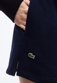 Χέρι που ξεκουράζετα ι στην τσέπη ενδύματος σε ναυτικό μπλε χρώμα με μικρό πράσινο λογότυπο κροκοδείλου κοντά στο τελείωμα.