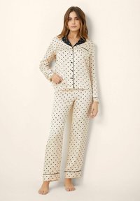 Polka dot pajama set in cream satin with black accents. Long-sleeve shirt features a notch collar; pants have an elastic waistband.