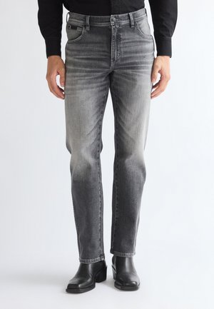 Homme portant un jean gris délavé, une chemise noire et des bottines en cuir noir, debout avec les mains détendues le long du corps.