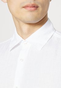 Camicia bianca in lino con colletto button-down, caratterizzata da un tessuto con texture e classici bottini, che mostra un design pulito e semplice.