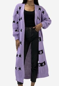 Liliowy sweter na drutach z otwartym przodem, ozdobiony czarnymi wzorami gwiazd. Długie rękawy i luźny fason, zestawiony z czarnymi jeansami.