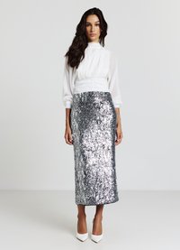 Blusa bianca con collo alto e maniche a sbuffo abbinata a una gonna a tubino argento con paillettes, che termina sopra la caviglia, indossata con tacchi bianchi.