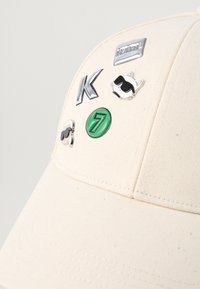 Gorra de béisbol beige con tejido texturizado adornada con varias chapas de metal y esmalte que presentan letras y un mapache con gafas de sol.