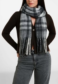 Donna che indossa un cardigan marrone scuro con bottoni, jeans grigi a vita alta e sciarpa grigia a quadri con estremità frangiate avvolta intorno al collo.