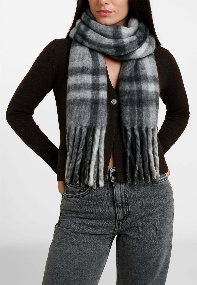 Donna che indossa un cardigan marrone scuro con bottoni, jeans grigi a vita alta e sciarpa grigia a quadri con estremità frangiate avvolta intorno al collo.