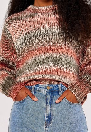 Personne portant un pull en tricot multicolore dans des tons roses et gris, les mains dans les poches d'un jean bleu taille haute sur fond blanc.