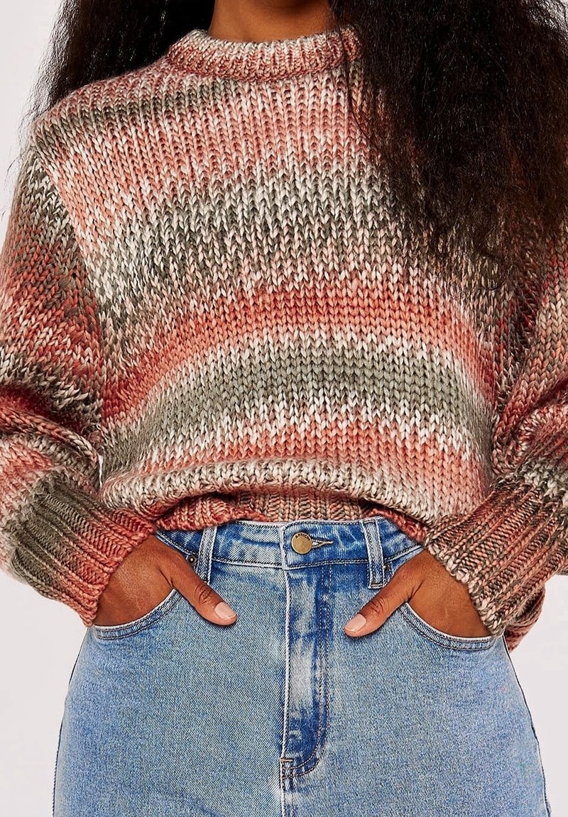 Personne portant un pull en tricot multicolore dans des tons roses et gris, les mains dans les poches d'un jean bleu taille haute sur fond blanc.
