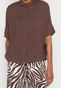 Bruine blouse met korte mouwen en knopen, met een gladde textuur en een relaxte pasvorm. Gecombineerd met witte broek met een bruin zebraprintpatroon.