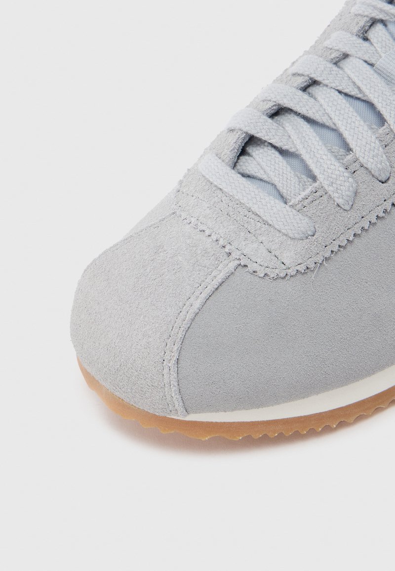 Suede sneaker in light gray with textured finish. Features flat gray laces, a rubber gum sole, and stitched accent detail on the upper.