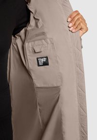 Hand die een open beige jas vasthoudt en een binnenzak laat zien met een zwart label waarop "Khujo™ original for a few" staat, tegen een lichte achtergrond.