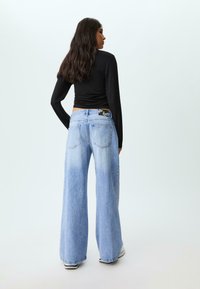 Donna con lunghi capelli scuri che indossa un top aderente nero a maniche lunghe e jeans a vita alta azzurri e larghi, in piedi con la schiena rivolta.
