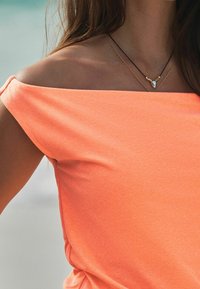 Oranssi, pehmeästä kankaasta oleva olkapäät paljastava paita lyhyillä hihoilla. Kaksi kerroksista kaulaketjua: yksi kultaisilla yksityiskohdilla ja toinen pisaramaisella riipuksella.