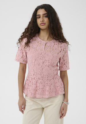 Młoda kobieta o długich kręconych włosach, ubrana w różową koronkową bluzkę z krótkim rękawem i jasnobeżowe spodnie na jednolitym tle.