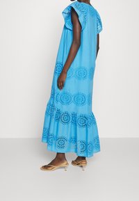 Robe longue en coton bleu avec broderie œillets et ourlet à volants. Manches avec détails découpés. Associée à des sandales à talons de couleur claire.