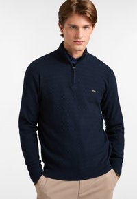 Suéter azul marino con cuello de cremallera, hecho de un tejido de punto texturizado, que presenta un sutil patrón en zigzag y un logo dorado en el lado izquierdo del pecho.