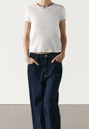 Personne portant un haut en maille blanc à manches courtes avec une petite bordure noire et un jean large foncé, les mains dans les poches.