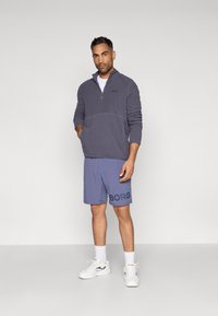 Γκρι φλις μισό-φέρετρο pullover με τσέπη καγκουρό, σε συνδυασμό με μπλε σορτς που διαθέτουν μεγάλη μαύρη εκτύπωση "BORG", φορεμένο με λευκά αθλητικά παπούτσια.